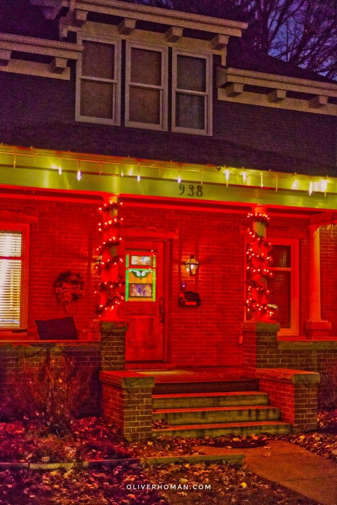 Red light from Christmas lights bathes the front porch of a brick house. White lights frame the roof of the porch from above. A stained glass insert in the front door is lit up from inside.