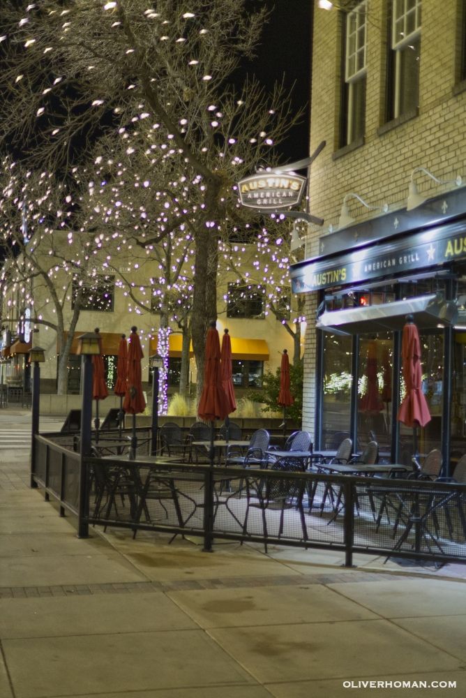 The patio of Austin's American Grill is lit up by surrounding trees and holiday lights in Old Town, Fort Collins, Colorado. Metal patio chairs and tables with red umbrellas are visible behind a metal fence. A concrete sidewalk made up of large squares divided by lines makes up the foreground of the image.