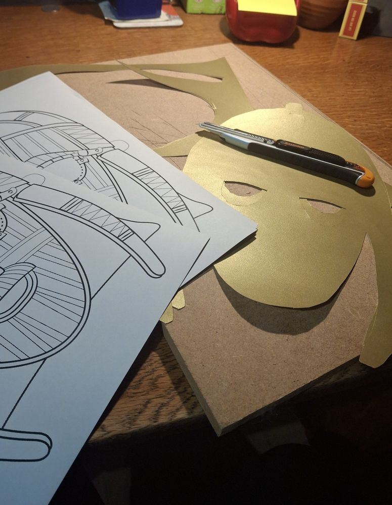 A pile of masks based on the Sutton Hoo helmet sit on a cutting board alongside a craft knife.