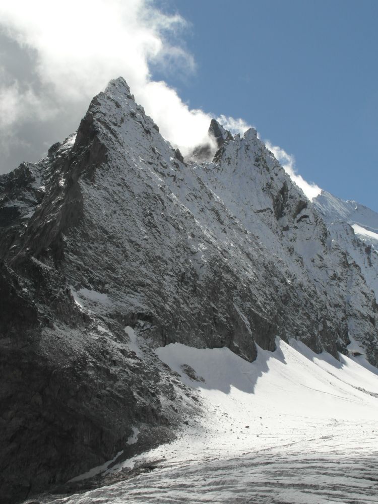 A mountain ridge dusted in snow above a snow curtain and glacier. Clouds 'steam' off the back side of the ridge. 