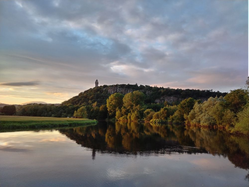 Tower on a hill above hills reflected in slightly ruffled river water. 