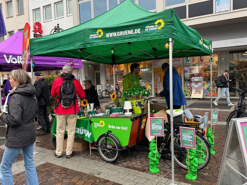 Fotografie eines Wahlkampfstands der brühler Grünen. Man sieht unter dem grünen Pavillon mit den Logos von Bündnis90 / Die Grünen unser grünes Lastenfahrrad mit den Wahlkampfgeschenken, wie zum Beispiel den grünen Windrädern. Unter dem Pavillon und davor stehen Menschen. Auf der rechten Seite sieht man den lila farbenen Pavillon einer anderen Partei, im Hintergrund die Schaufensterfront einer Ladenzeile. Vor dem Stand stehen ausserdem grüne Gartenzwerge mit Schildern in der Hand. Darauf die Slogans "Damit Brühl ein Zuhause für alle bleibt" und "Damit sich Firmen gerne hier niederlassen".