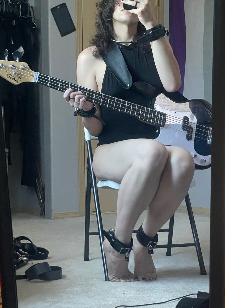 Mux sits in a chair wearing a bodysuit and locked leather cuffs while holding a bass guitar 