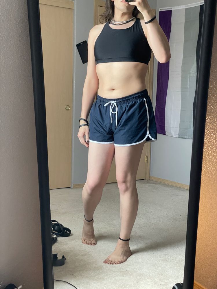 Mux stands wearing a black swimsuit top and white lined swim shorts along with a metal band collar and silicone band wrist/anklets 