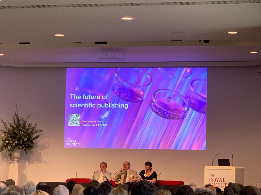 Three white people sitting on a stage (Bill Sutherland, Mark Walport, and Sophie Meekings) with a Royal Society branded lectern in one corner and a slide projected behind them entitled "The future of scientific publishing"