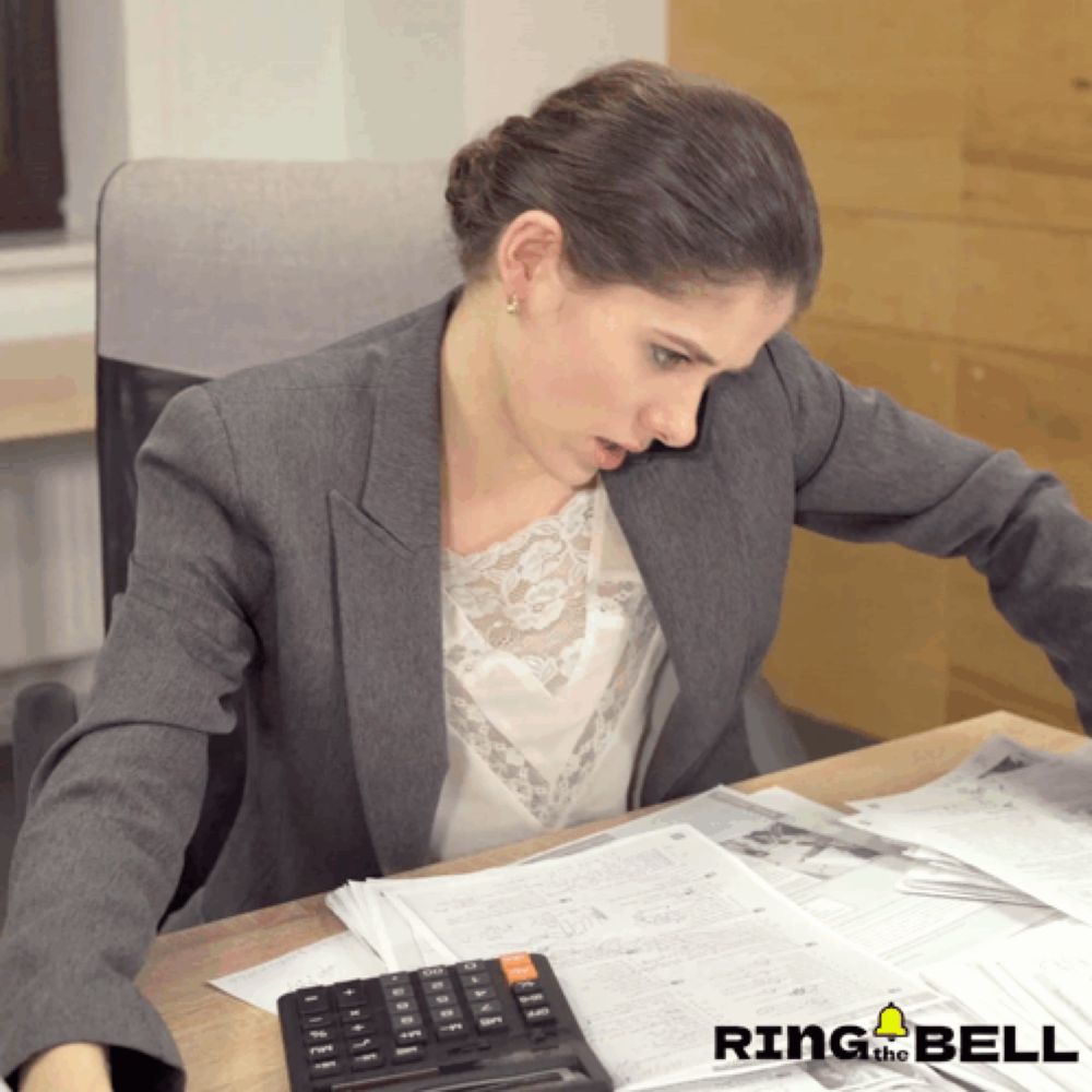 a woman sits at a desk with papers and a calculator with ring and bell in the corner