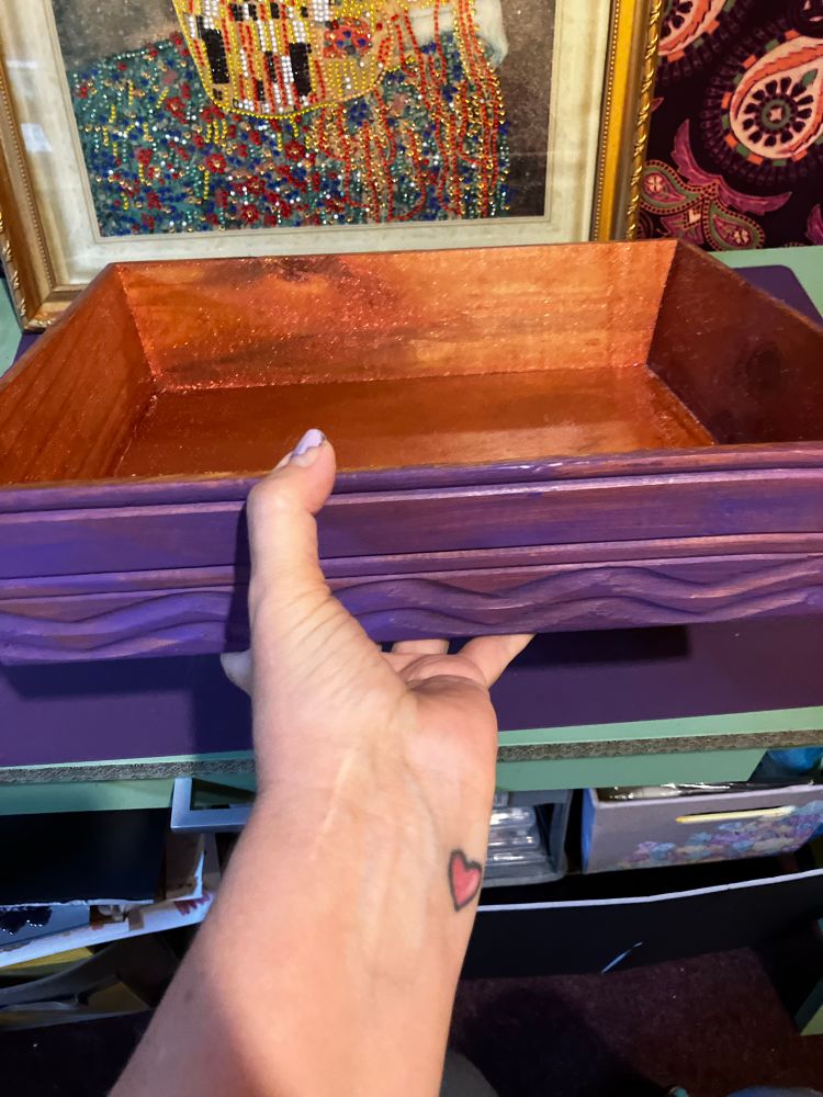 Woman’s hand holding a wooden tray with sides. The interior is painted metallic gold, and the outside is bright purple. 