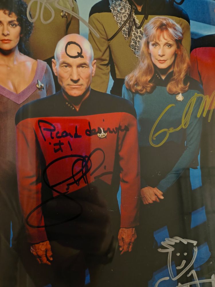 A cropped photo of the cast of Star Trek The Next Generation. There are parts of other characters and autographs visible in the image, but front and center is Captain Picard. Written on his forehead in black sharpie is the letter Q. Across his chest is written 'Picard deserves it', and underneath that is the signature of John de Lancie.