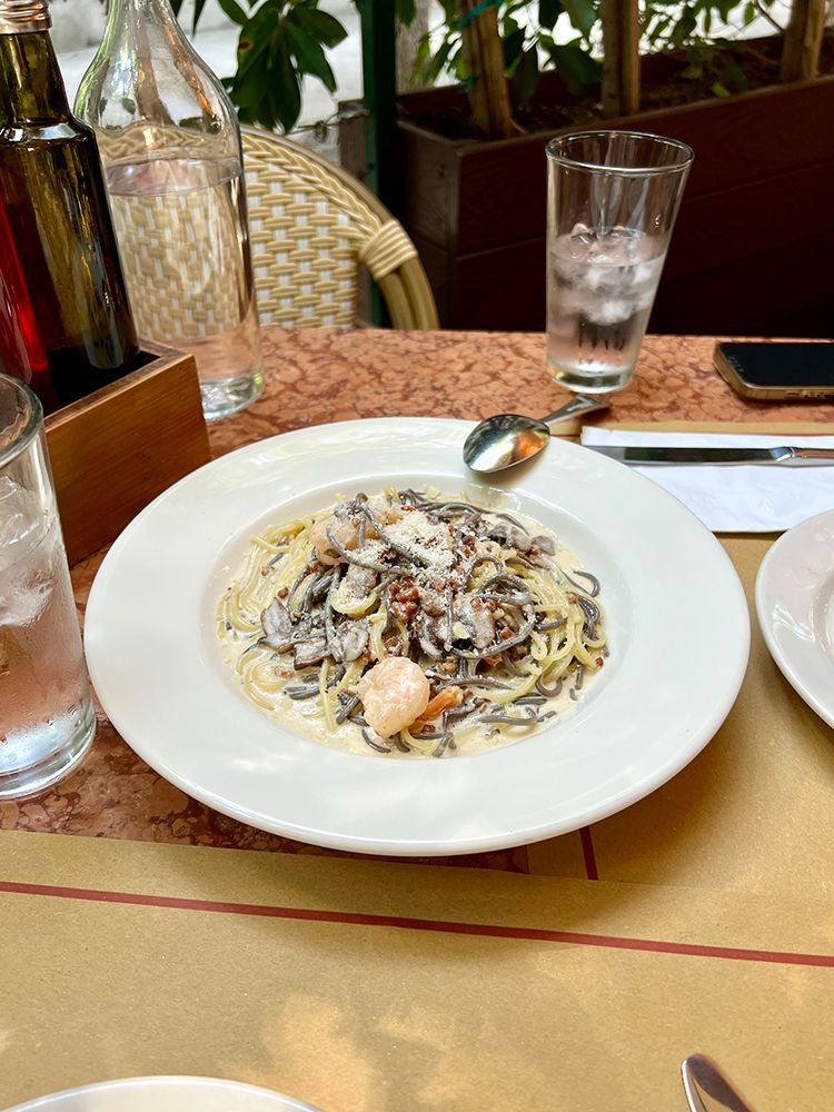 An outdoor dining table in the style of a bisto. At the center of the table is a plate of black and white vermicelli, shrimp, wild mushroom, and roasted pancetta, tossed in a creamy bisque.