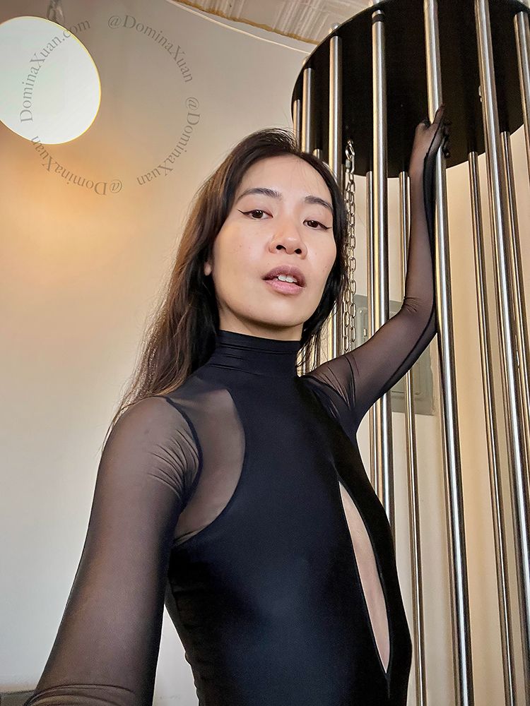 An Asian femme with long black hair, wearing a black Anoeses bodysuit. They are standing next to a vertical cage with their left hand grasping the top of one of its metal bars.