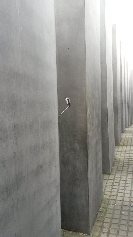Man sieht eine Reihe der übermannshohen grauen Beton-Stelen, aus denen das Mahnmal besteht. Der Boden ist gepflastert. 

Zwischen den ersten beiden Säulenreihen ragt ein Selfiestick in den Gang.