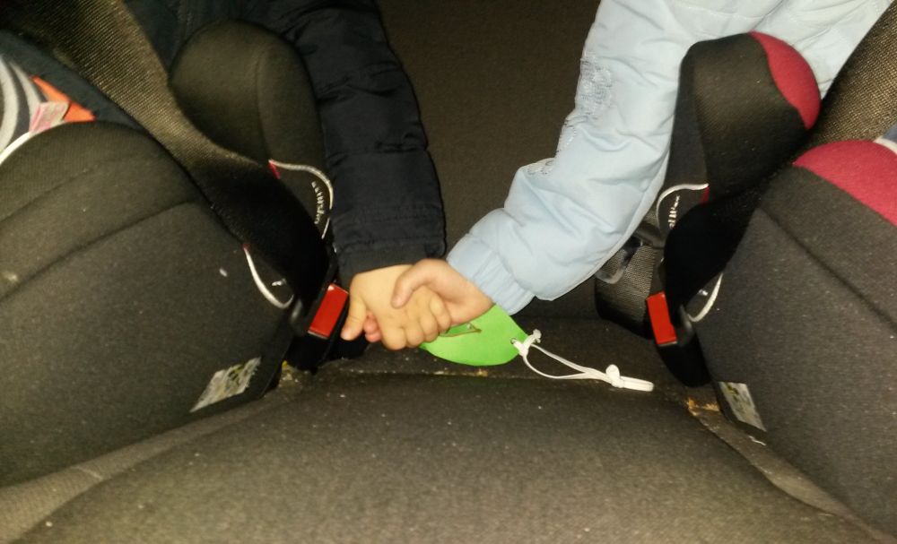 Man sieht den unteren Teil von zwei Kindersitzen in einem Auto. Darin sitzt jeweils ein Kind, man sieht aber nur ein kleines Stück Hüfte, den Gurt und zwei Arme, die rechte Hand des linken Kindes und die linke Hand des rechten Kindes halten einander fest. Das eine Kind trägt eine hellblaue, das andere eine dunkle Jacke. 

Zwischen den beiden Sitzen hinter den Händen liegt eine selbstgebastelte hellgrüne Maske aus Tonpapier mit Hosengummi zur Befestigung.  