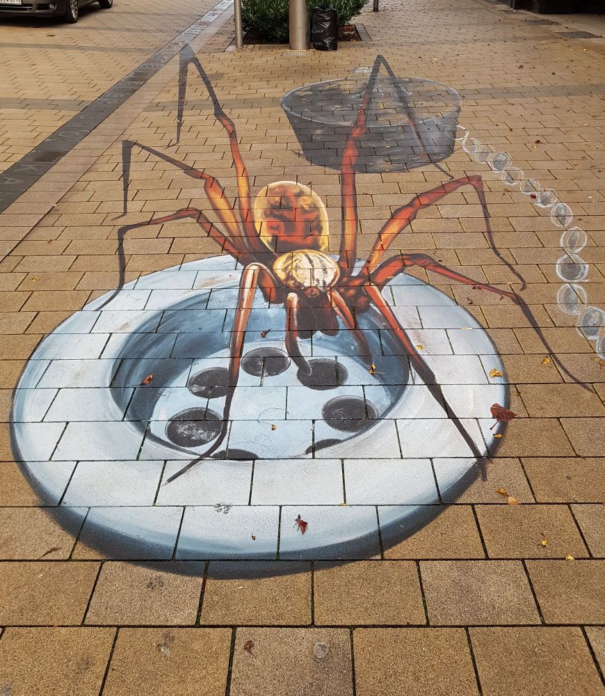 Auf dem Bild sieht man das gelbe Pflaster einer Fußgängerzone. 
Darauf hat ein*e Künstler*in mit Kreide eine riesige orangefarbene Spinne gemalt, die auf einem überdimensionierten Abfluss hockt. Rechts hinter ihr liegt an einer Kette der ebenfalls übergroße Stöpsel. 