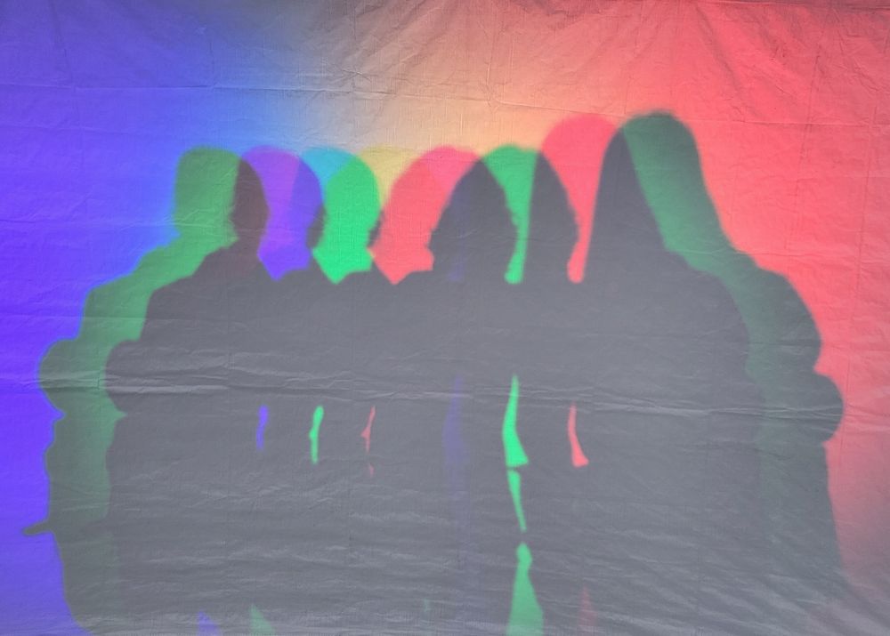 Auf dem Bild sieht man die Schatten von drei Personen auf einer Leinwand. Es gibt (von links nach rechts) blaue, grüne und rote Strahler, so dass dadurch zusätzlich zum dunklen Kernschatten seitlich leicht versetzte Halbschatten entstehen. Die drei Personen stehen in einer Reihe und halten sich im Arm. Die beiden linken Personen sind etwa gleich groß, die rechte Person ist etwas größer. 