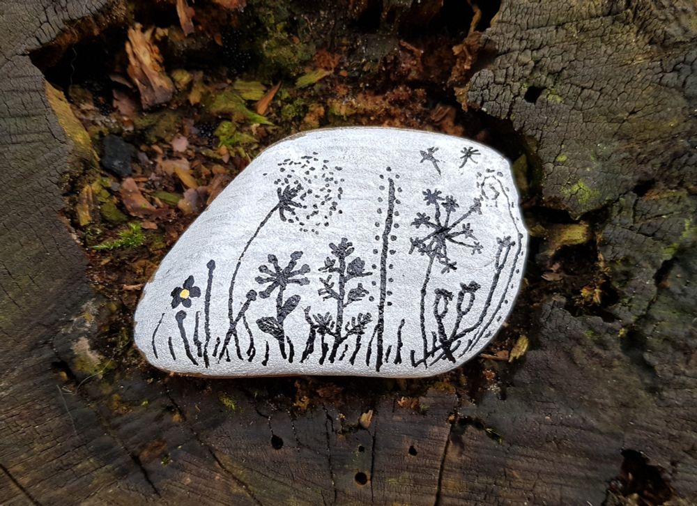 Auf einem Baumstumpf liegt ein weißer Kieselstein. Er ist nicht ganz rund, unten ist er abgeflacht und von links unten geht er sehr schräg nach links oben.

Der Stein ist mit einem schwarzen Stift bemalt, man erkennt verschiedene Gräser, Blüten, eine Pusteblume.