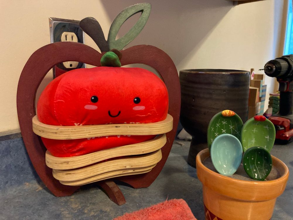 An oversized anthropomorphic plush smiling apple squeezed into a handmade wooden apple holder. There are also some ceramic spoons that look like cactus 