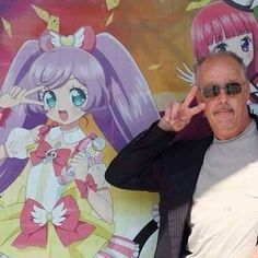 A picture of a middle-aged man wearing sunglasses and posing in front of a print of an anime-style idol. He is recreating her pose by raising a peace sign close to his eyes.