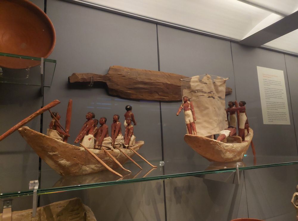 Two wooden boats, staffed with wooden oarsmen with tiny oars. One of the boats has a linen sail.