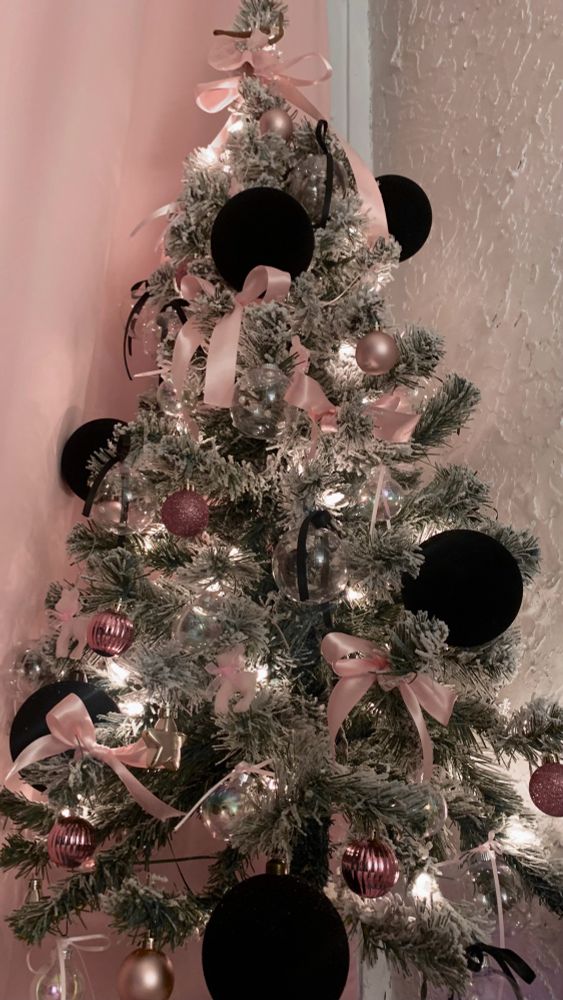 Green flocked mini Christmas tree covered in black and pink ornaments, baby pink bows and black ribbons. Sitting on a white stand in front of white wall and pink curtains.