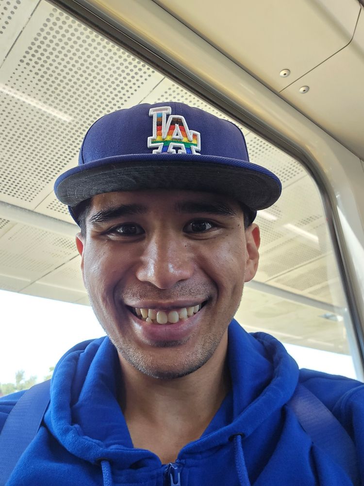 sirhapless poses for selfie inside Los Angeles light rail car wearing blue hoodie and Los Angeles Dodgers cap with "L A" in progress pride colors