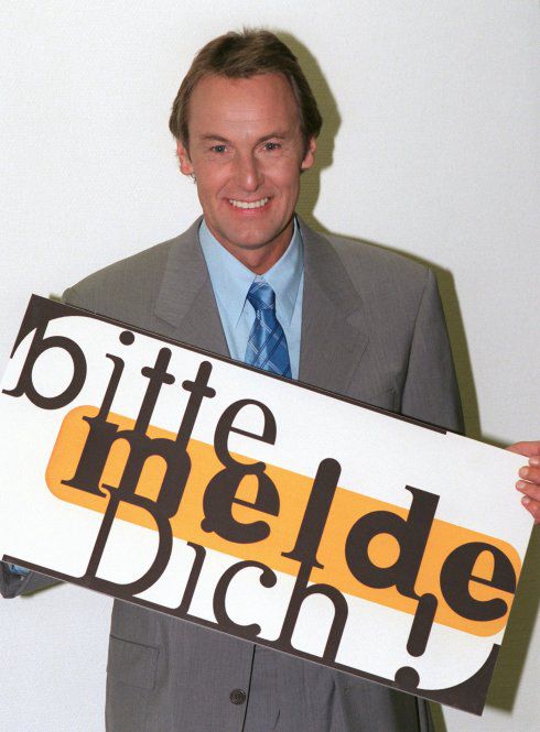 junger jörg wontorra in grauem anzug mit helblauem hemd und krawatte in etwas dunkleren blau. Er hält ein schild mit der aufschrift „bitte melde dich!“ vor seinem körper