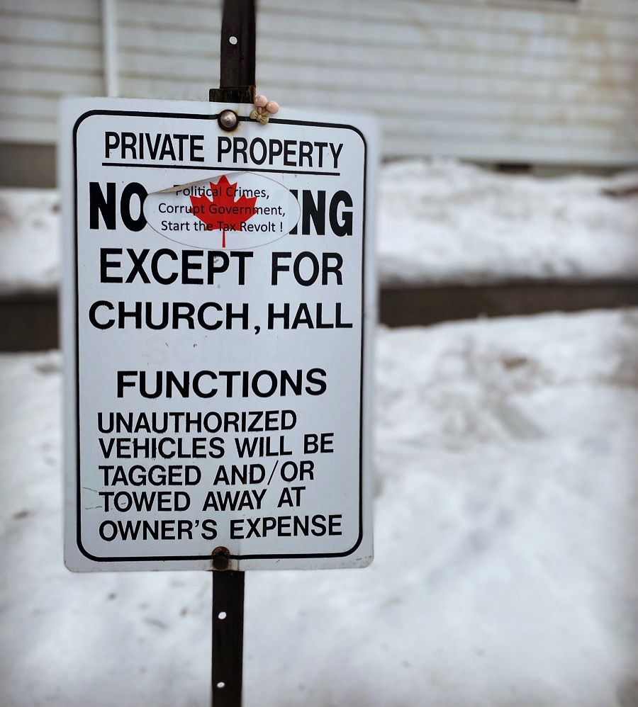 A picture of a white sign reading:
 “PRIVATE PROPERTY
NO PARKING EXCEPT FOR CHURCH, HALL FUNCTIONS
UNAUTHORIZED VEHICLES WILL BE TAGGED AND/OR TOWED AWAY AT OWNER’S EXPENSE” 
with a white oval sticker with a red maple leaf in the centre covering the PARKI of PARKING that reads: “Political Crimes, Corrupt Government, Start the Tax Revolt!” in the foreground with a building with dingy white siding, sidewalk and snow covered ground in the background.