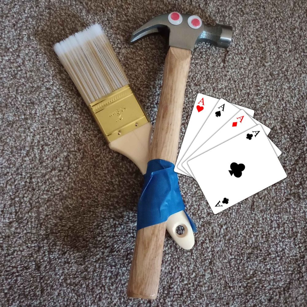 A Hammer with googly eyes taped to a paintbrush next to a set of Ace playing cards