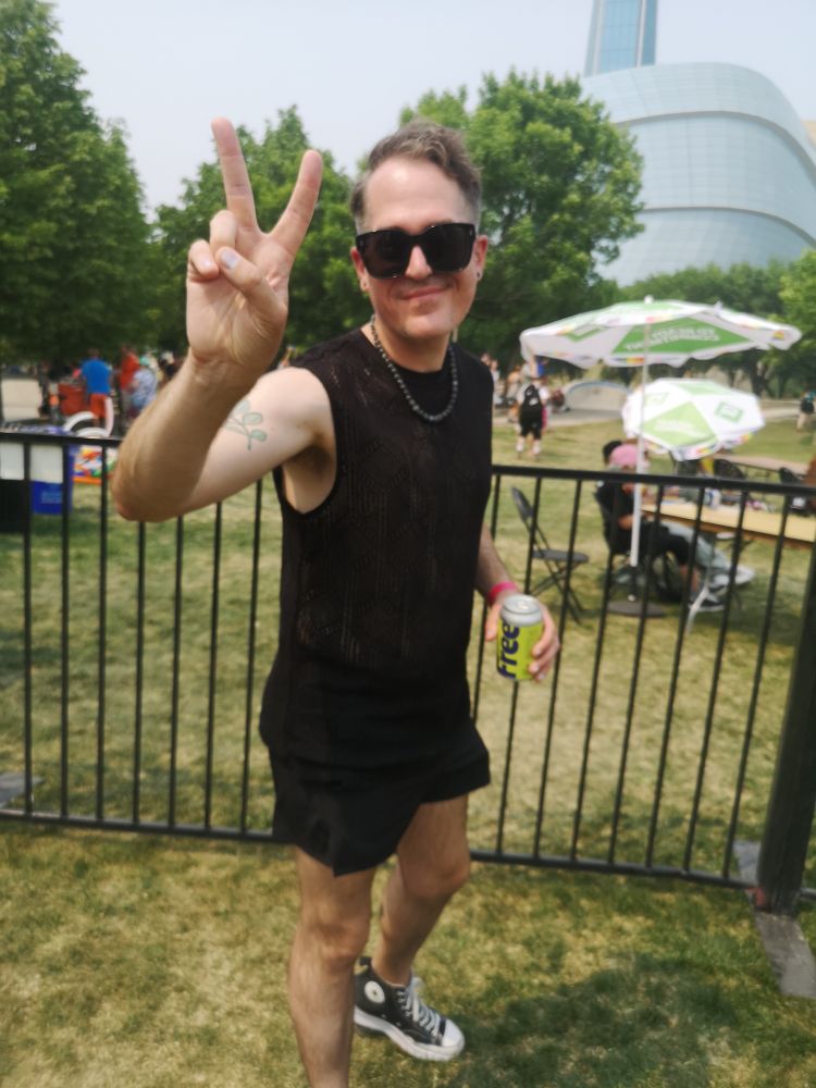 Me, at Winnipeg Pride (Sunday, 1 June 2025), flashing a peace sign whilst wearing all black & drinking a watermelon hibiscus sparkling water.