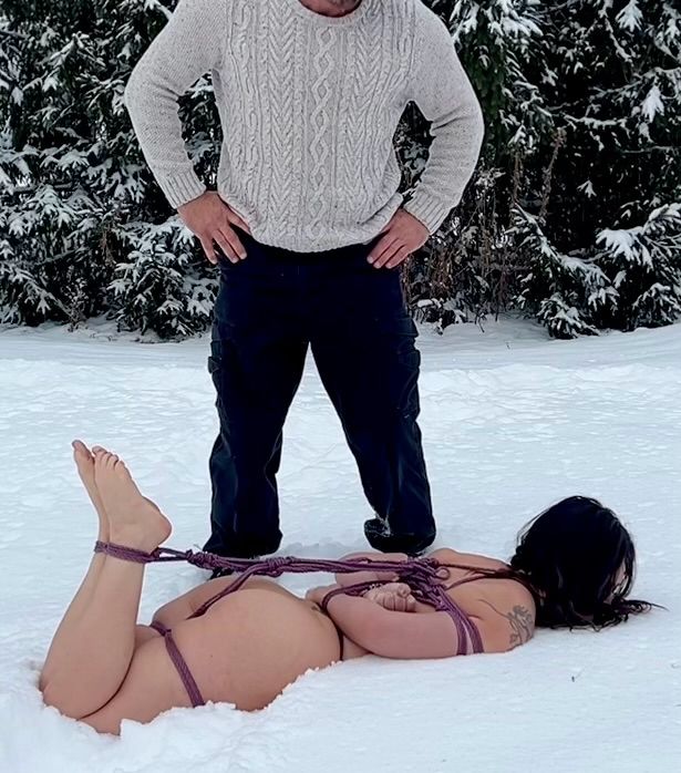 A man stands over a bound naked woman lying face down in the snow. 