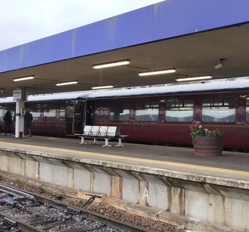 Dunkelroter Personenwagen mit iffenen Türen auf einem Bahnsteig, Schild "Fort William", gälischer Name unleserlich.