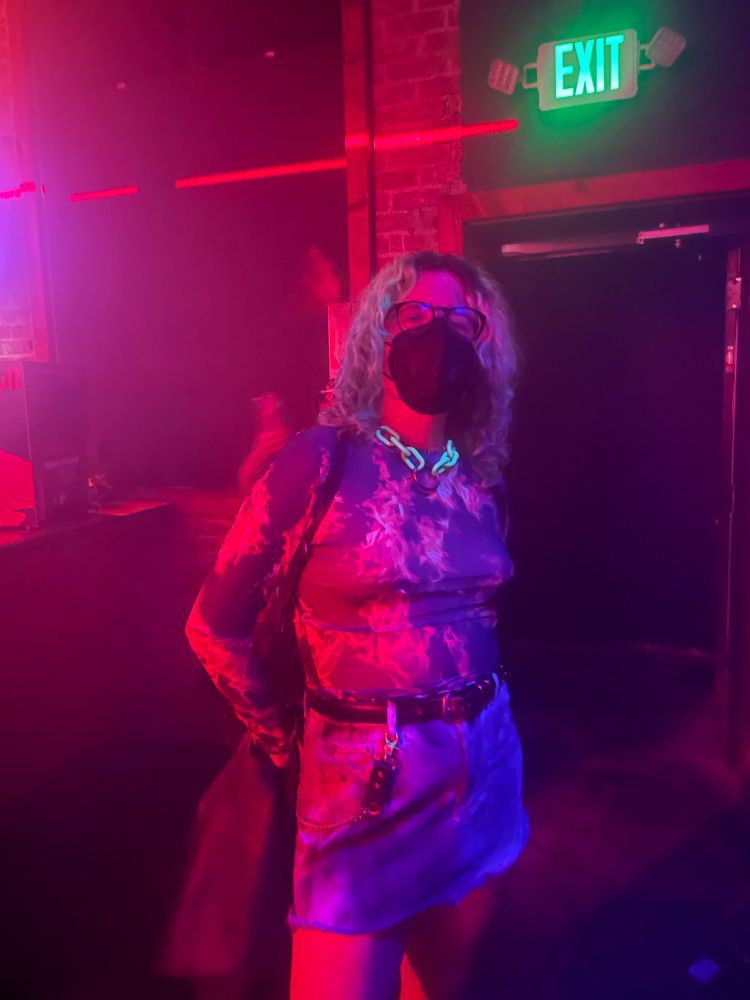trans girl in a black mask and flash top and skirt standing under red and blue lights in a club