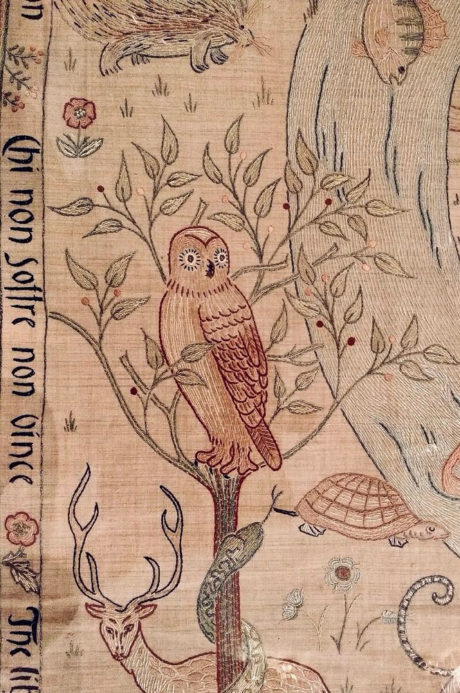 Embroidery depicts an owl in a tree, the trunk is wound by a snake climbing, there’s a deer at the lower portion of the trunk, there’s a tortoise to one side approaching a river that has a fish swimming downstream and a hedgehog at the very top, above the tree. There is writing down one side, but I don’t know what it says. 