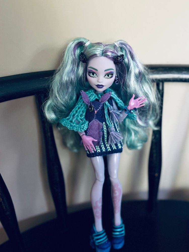 Monster High Twyla doll in a short knit dress and a knit cape, made of blue green yarn with purple tassels
