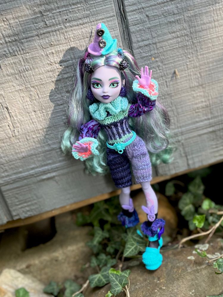 monster high twyla doll dressed as a clown. her hat is conical, half blue and half purple, sewn with felt. she wears knitted blue and purple ruffles on her neck, arms, and legs. she also has a knitted jumpsuit with vertical stripes, and black socks with purple felt bunny faces at the tops