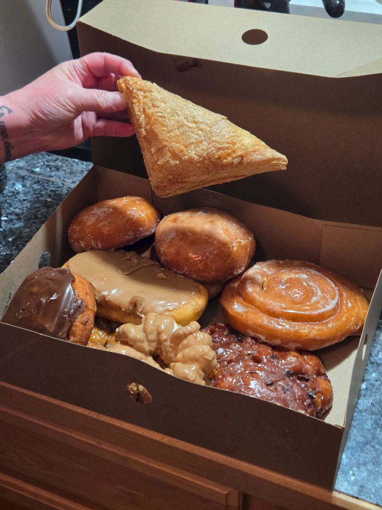 A box of donuts.  Someone is taking a cream cheese turnover. Many more remain. 