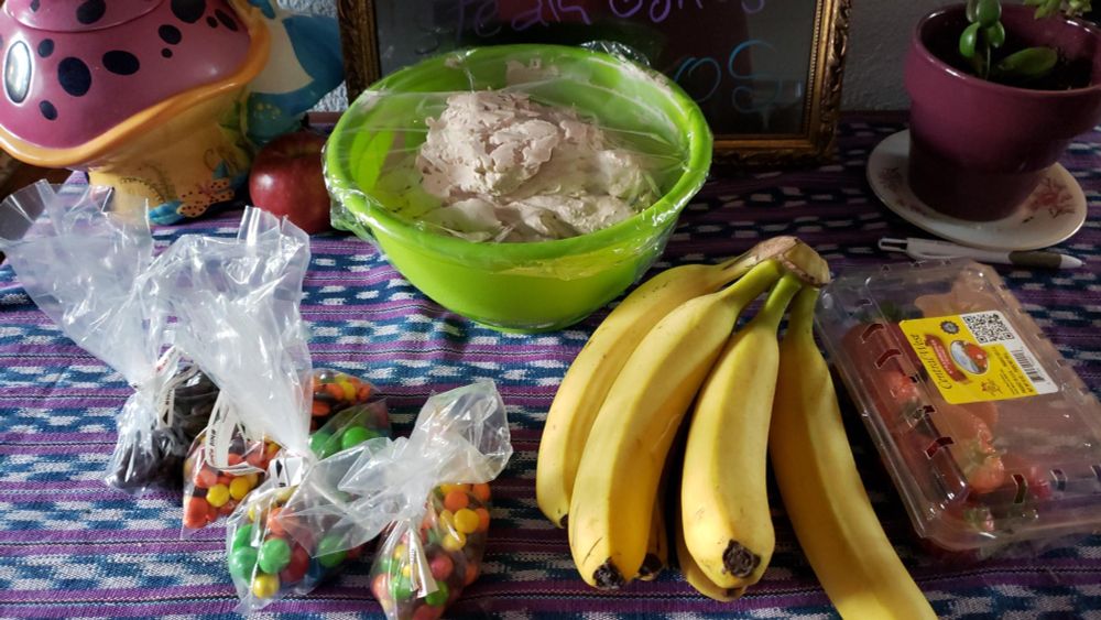 We are preparing for a banana split sunday Sunday!  This picture shows for bags of candy pieces (to be used as toppings), a bunch of bananas, a pack of strawberries, and a large bowl of homemade chocolate whipped cream.  These items sit upon a patterned tablecloth, between an Alice in Wonderland cookie jar and a potted jade plant. 