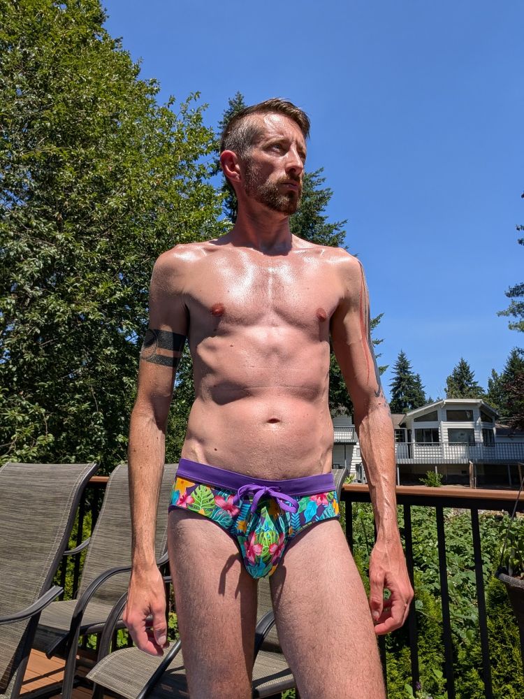 Very gay man wearing a brief-cut swimsuit on a deck and he is glowing with happy summer vibes