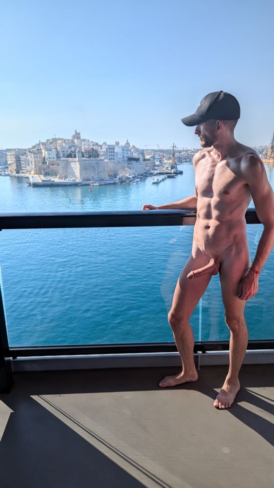 Naked man standing on a balcony overlooking the water and a coastal city. He's wearing a hat and feels good about it. 