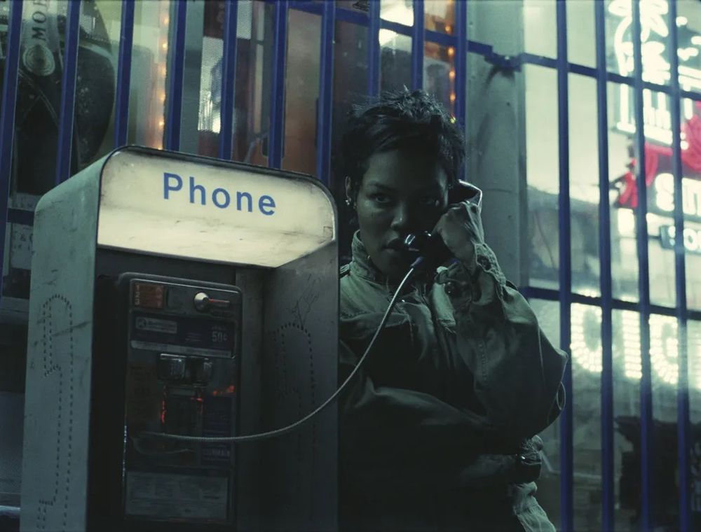 Teyana Taylor talking alone on a payphone outside of a gas station in the dark of the night with an intense, determined expression on her face in a scene from Paul Thomas Anderson's ONE BATTLE AFTER ANOTHER. 