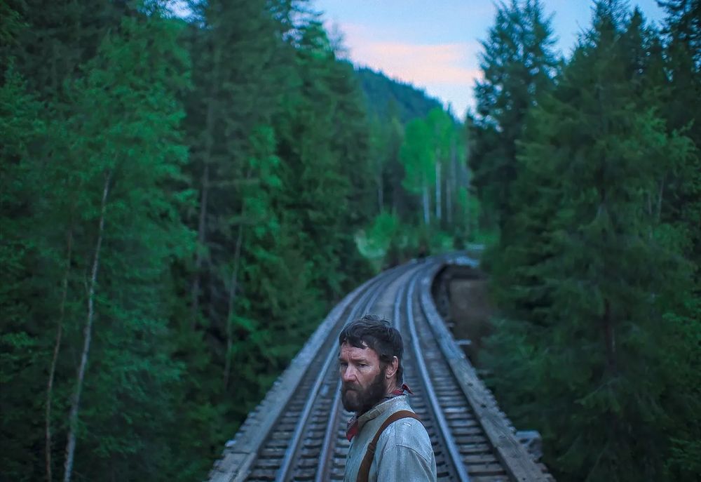 Joel Edgerton standing alone in the middle of a railroad as the vast and beautiful forests of Washington tower all around him in a still from TRAIN DREAMS. 