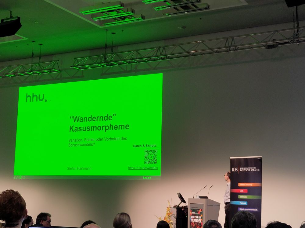 Foto: Ein Mann steht auf einer Bühne am Rednerpult. Im Hintergrund eine Powerpoint-Folie mit der Aufschrift ""Wandernde" Kasusmorpheme. Variation, Fehler oder Vorboten des Sprachwandels?" Der Folienhintergrund ist giftgrün.