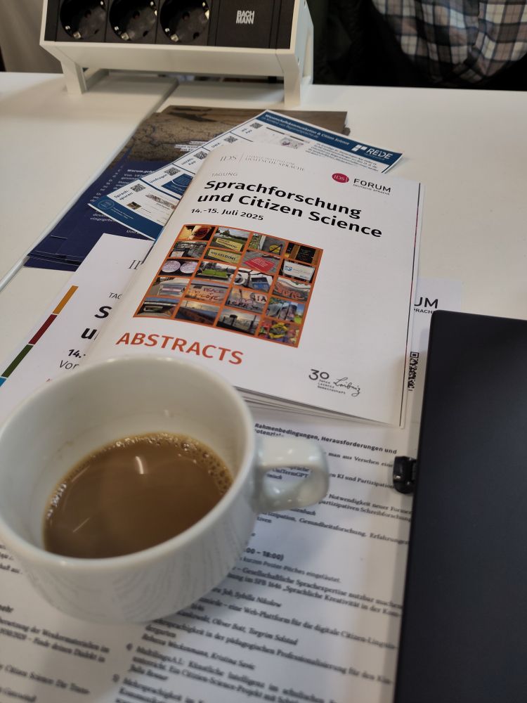 Foto des Abstracthefts der Tagung "Sprachforschung und Citizen Science" auf einem Tisch, daneben eine halbvolle Kaffeetasse.