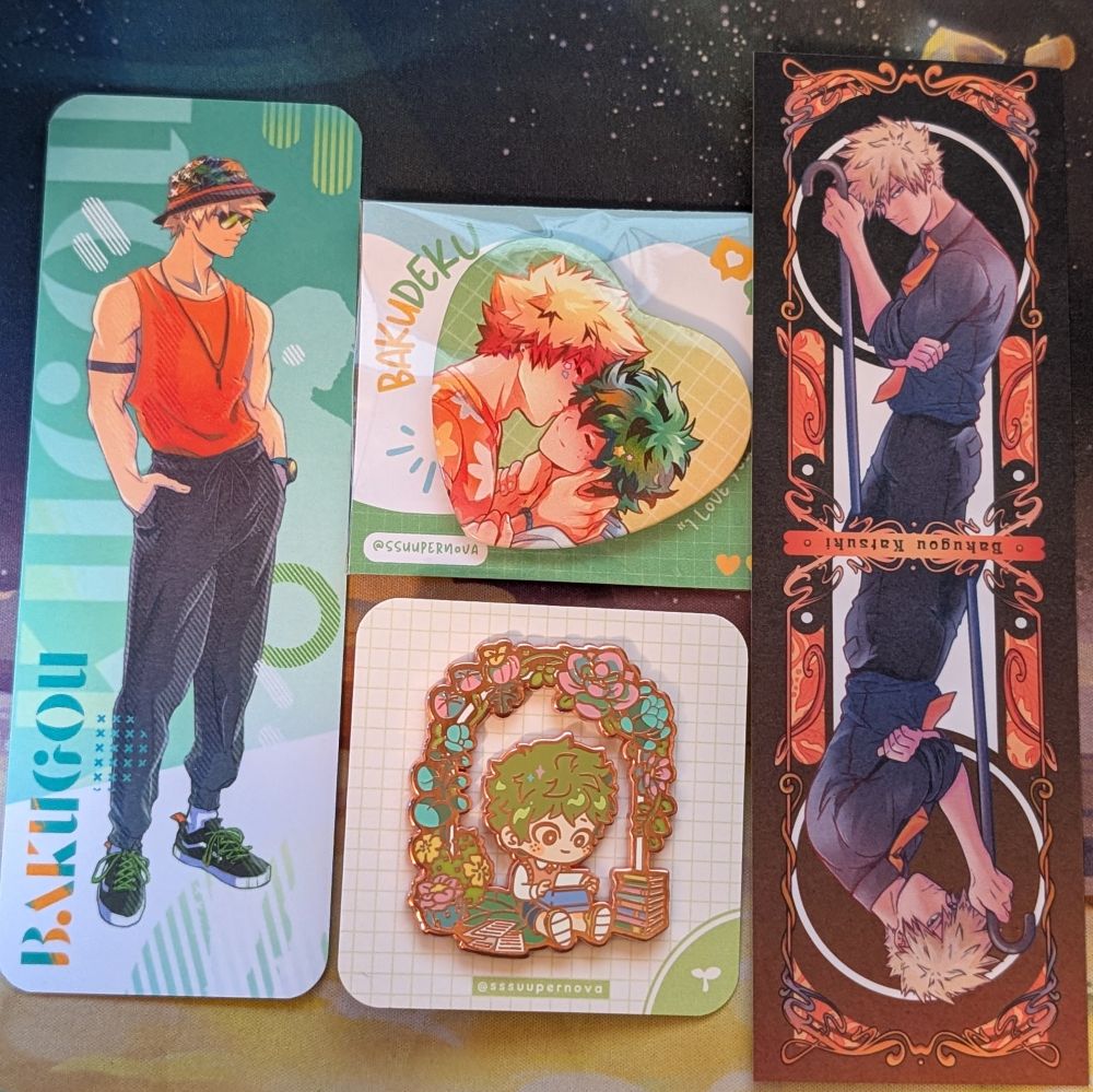 two bookmarks of bakugou katsuki, a heart-shaped button pin of katsuki kissing izuku's cheek, and an enamel pin of a young izuku using a typewriter while surrounded by books and flowers