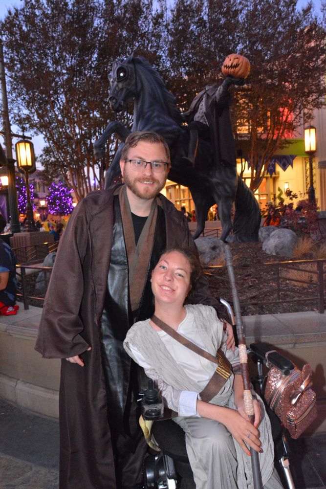 A white woman in a wheelchair wearing a Ray Star Wars costume with a white man standing behind her wearing a Jedi costume.