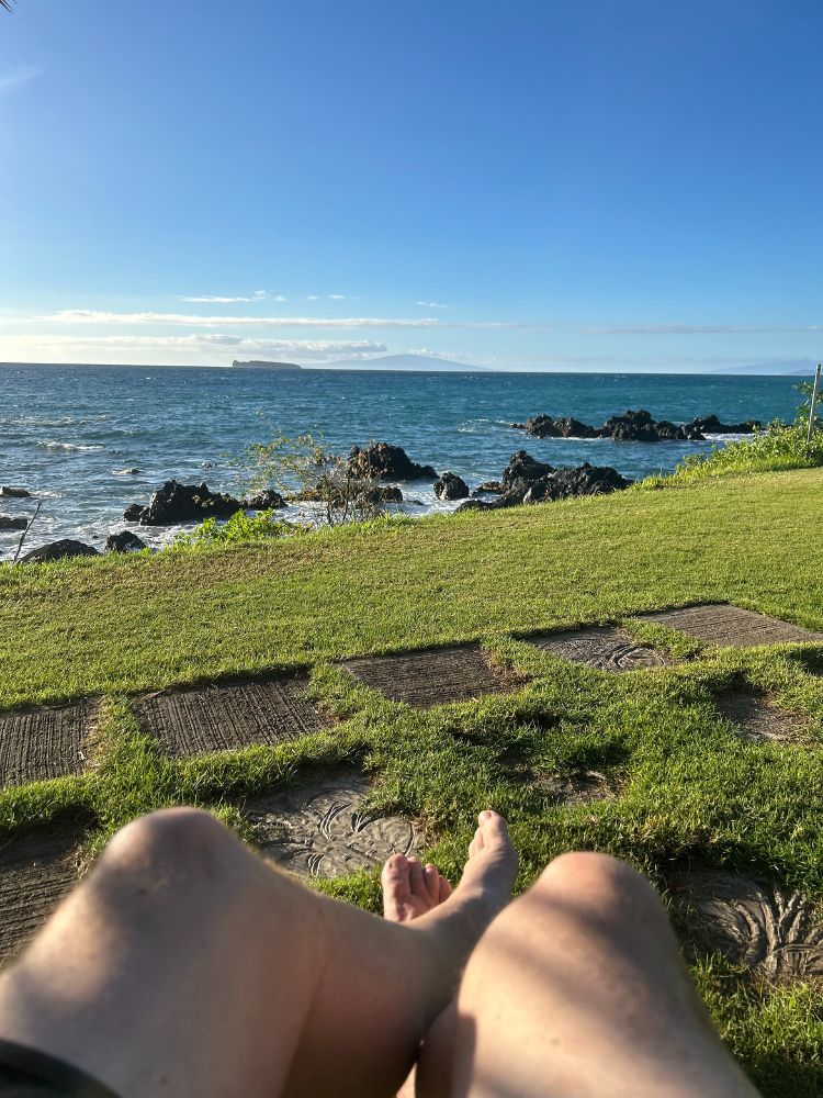 “View from a seated perspective on a grassy coastal lawn, with bare legs and feet in the foreground resting near stone stepping pads. Beyond the lawn, dark volcanic rocks line the shoreline as small waves roll in from a deep blue ocean. A low island and a larger mountainous landmass sit on the horizon beneath a clear, bright blue sky.”