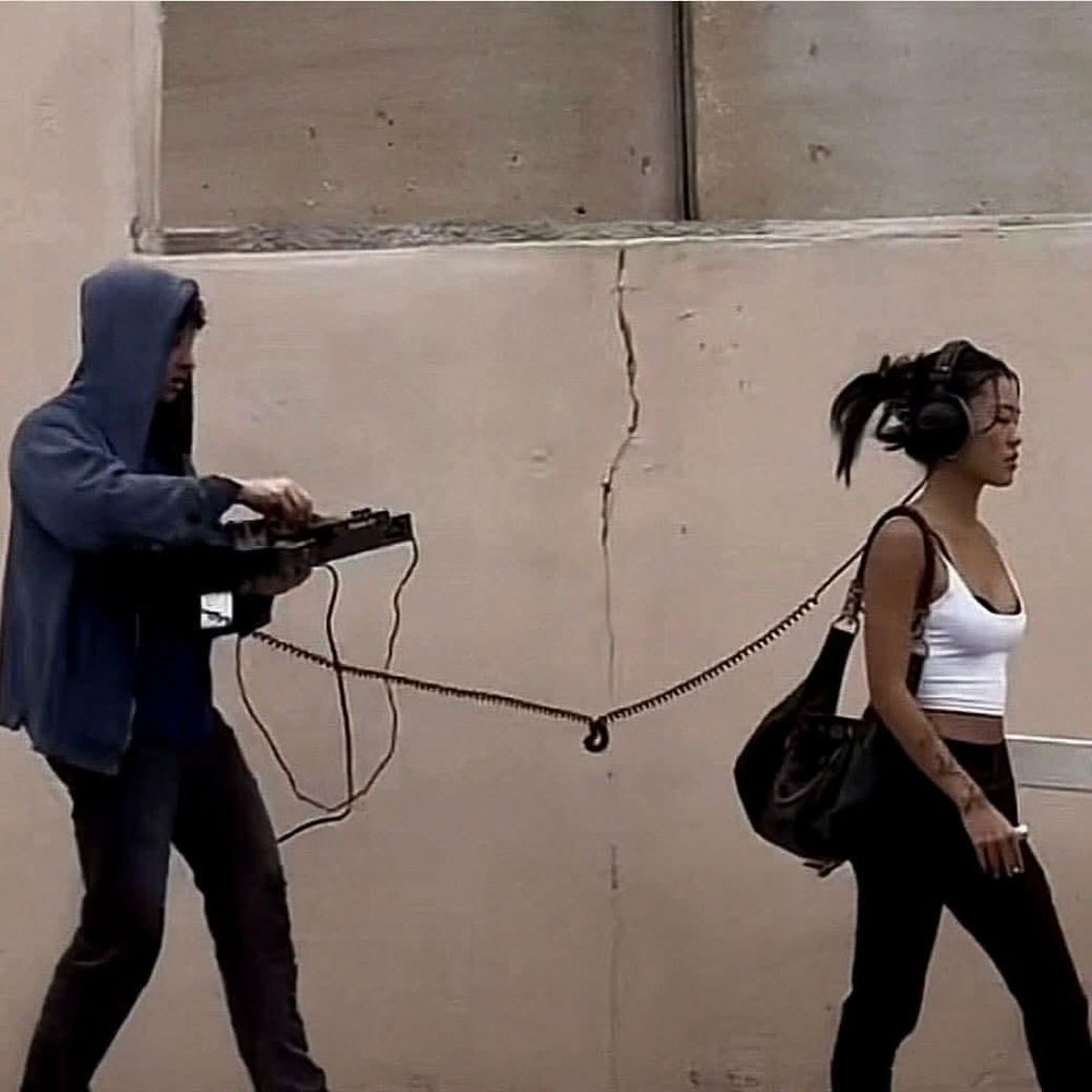 a girl walking with wired headphones on

right behind her, there's a dj mixing some music, with her headphone wire going straight to his controller