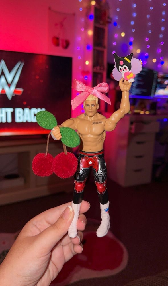 my hand holding up my cody rhodes action figure. he’s got a pink bow on his head, a kuromi in his hand + cherries in his other hand