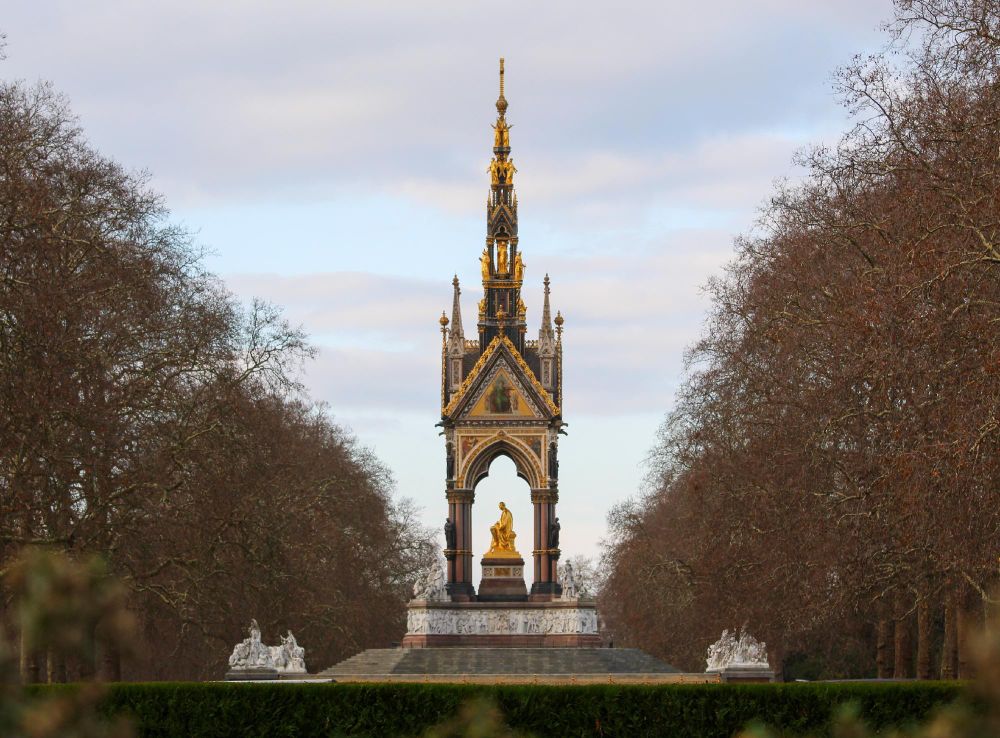 View of the Albert Memorial in Kensington Gardens, London. The memorial is framed by leafless trees and features an ornate golden spire atop a larger sculptural set with statues.