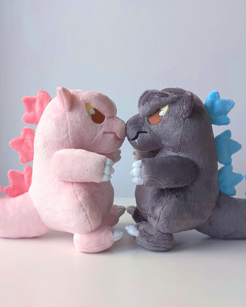 A photo of two Godzilla plushies facing each other with their noses touching. The one on the left is light pink with darker pink spikes on his back, the other is grey with blue spikes. 
