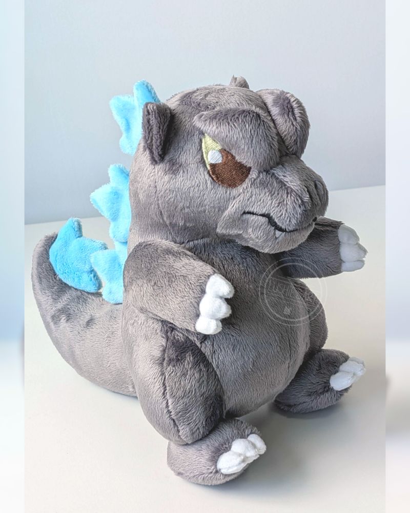 Photo of a Godzilla plushie. He is grey with blue spikes on his back.